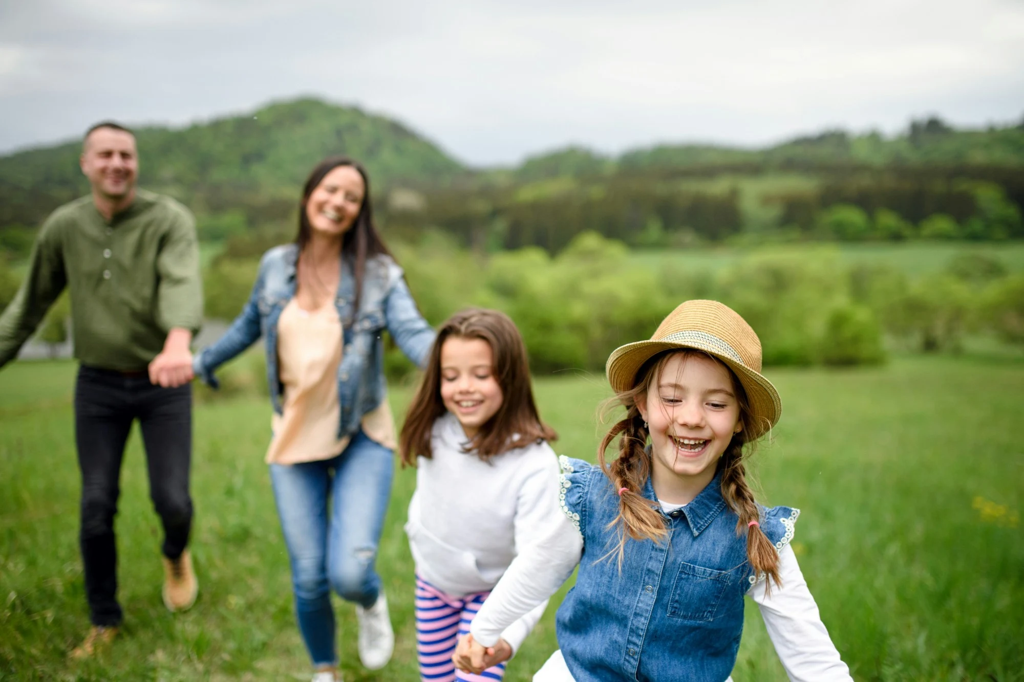 Parents and their two daughters running hand in hand through a green meadow.